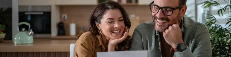 Happy mid adult couple sitting at a kitchen table, laughing while reviewing good insurance news 
