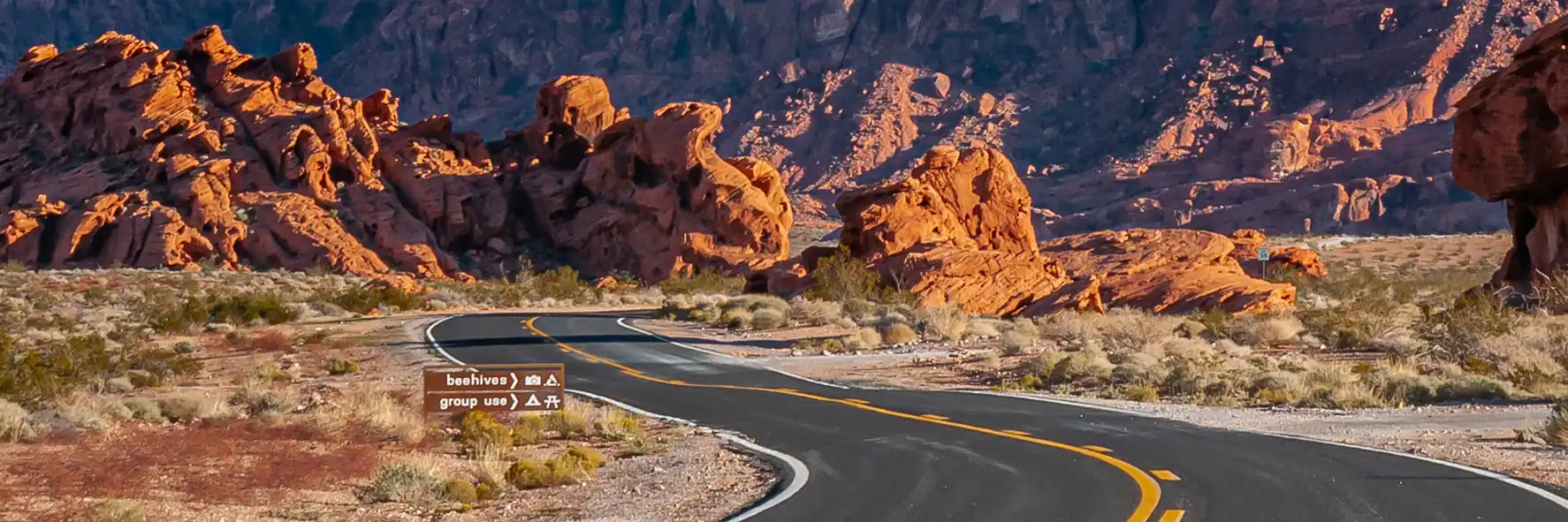 Sunset landscape under blue sky with black asphalt road meandering through dry desert with red colored mountains and rocks.