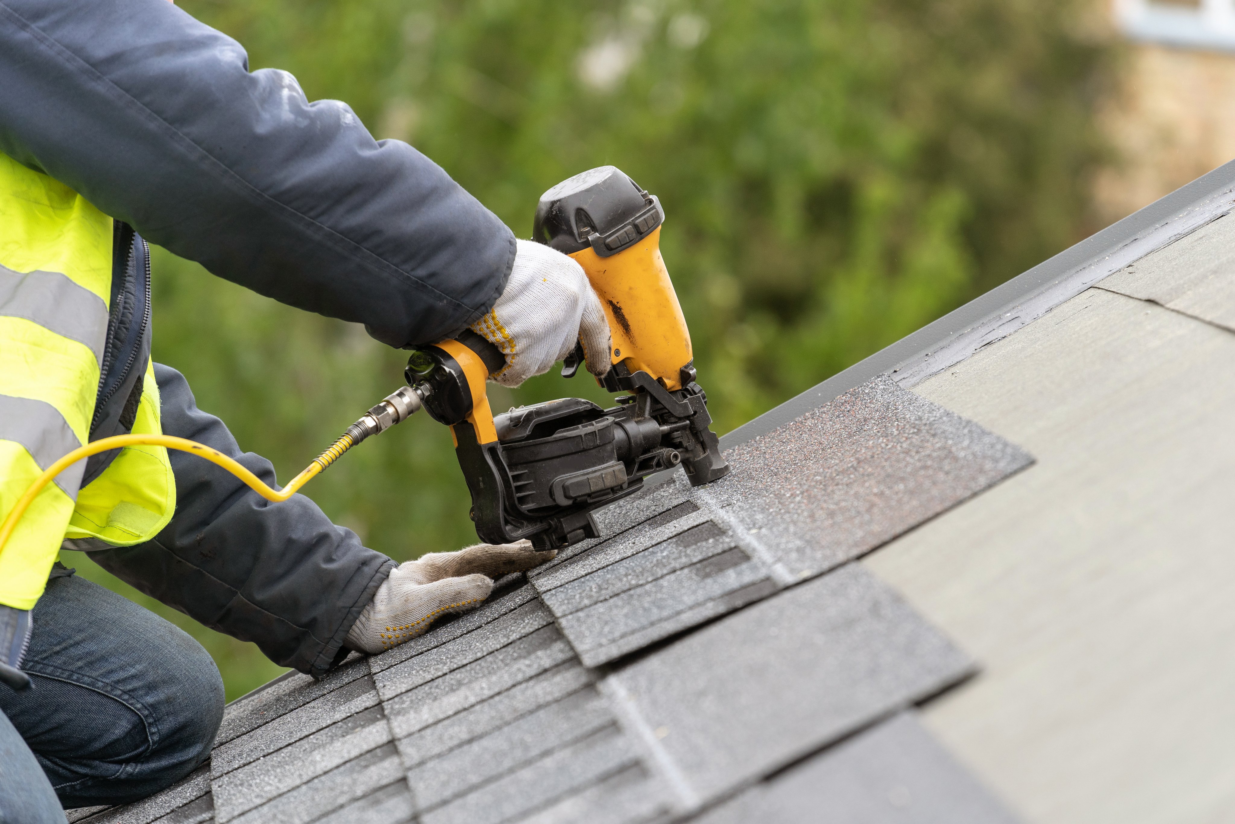Roofer using air or pneumatic nail gun and installing asphalt or bitumen tile on top of the roof