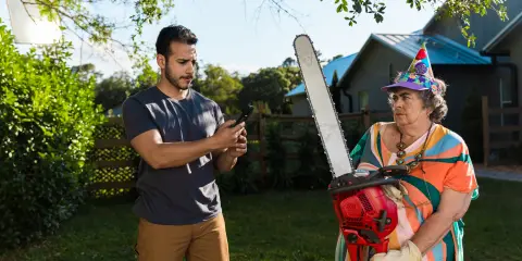 Man taking a photo of an older woman in a colorful outfit and party hat borrowing his chainsaw in a backyard