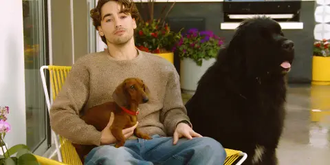 Man sitting on a patio chair holding a small dachshund while a large black dog sits beside him