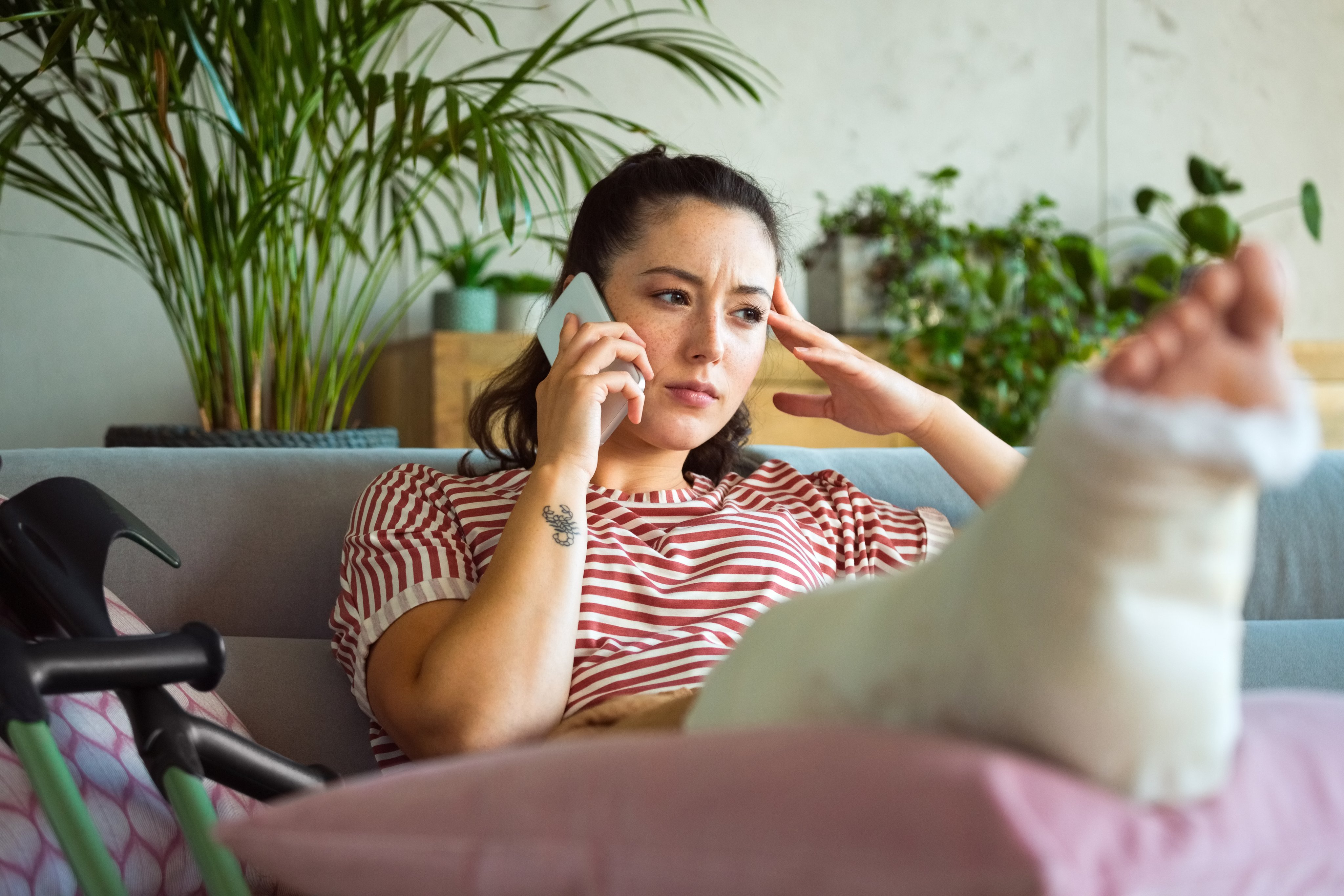 Worried young man with broken leg in plaster cast lying down on sofa at home and talking on smart phone