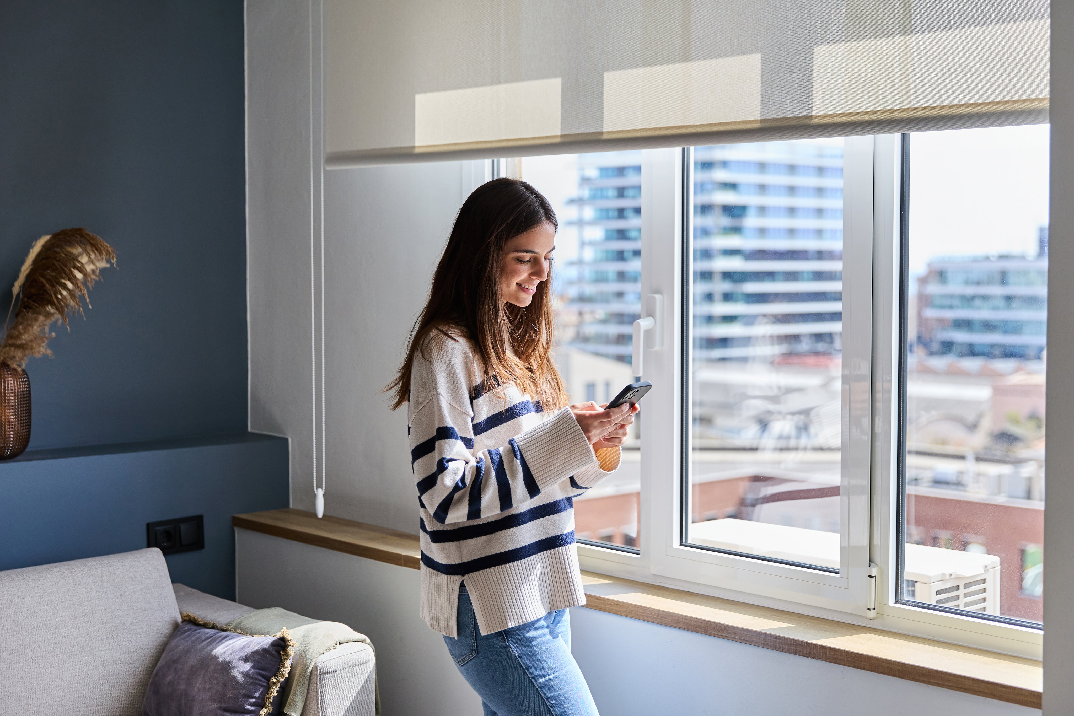 Young woman using smartphone at home standing by window in living room