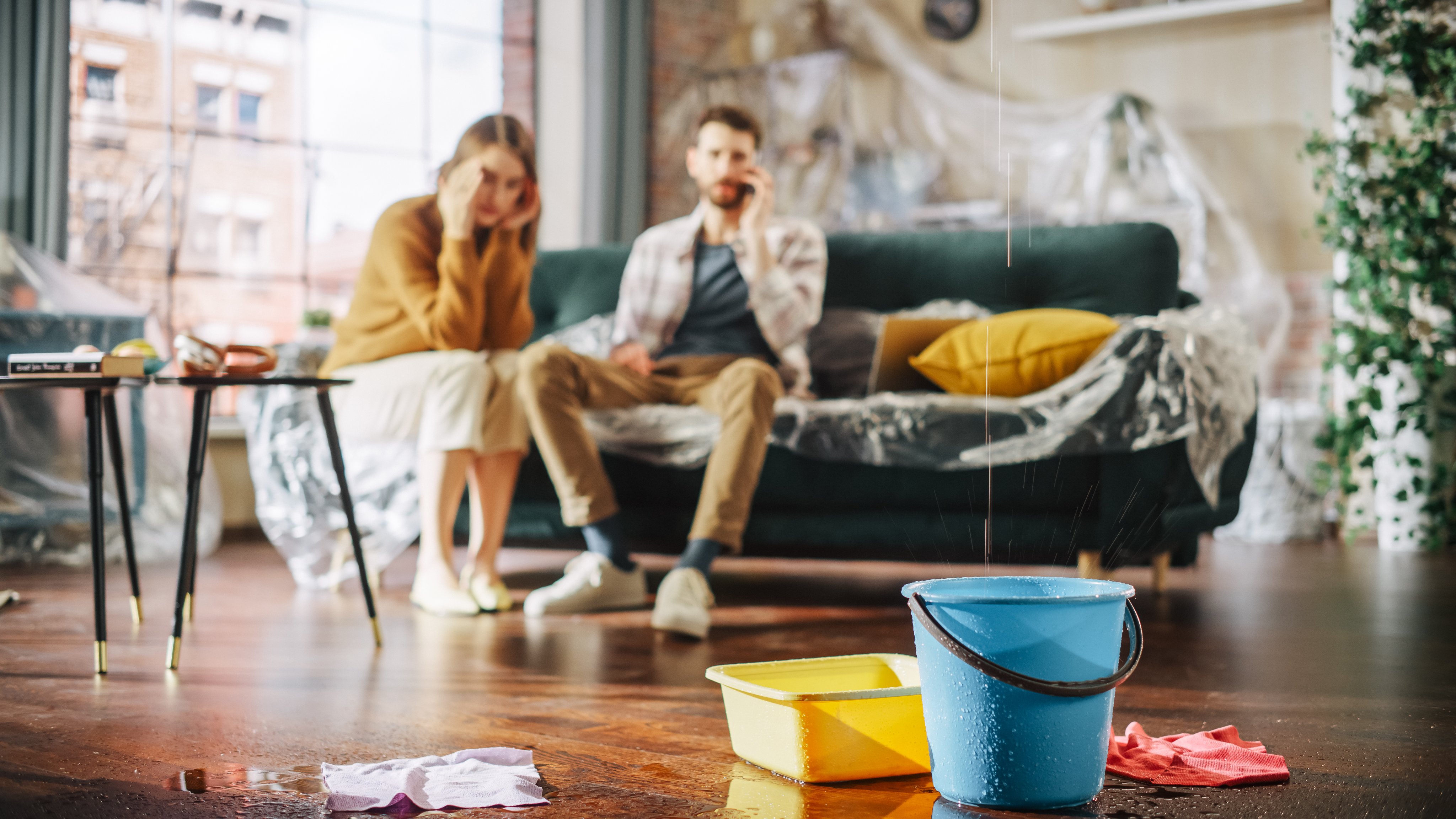 Panicing Couple In Despair Sitting on a Sofa Watching How Water Drips into Buckets in their Living Room.