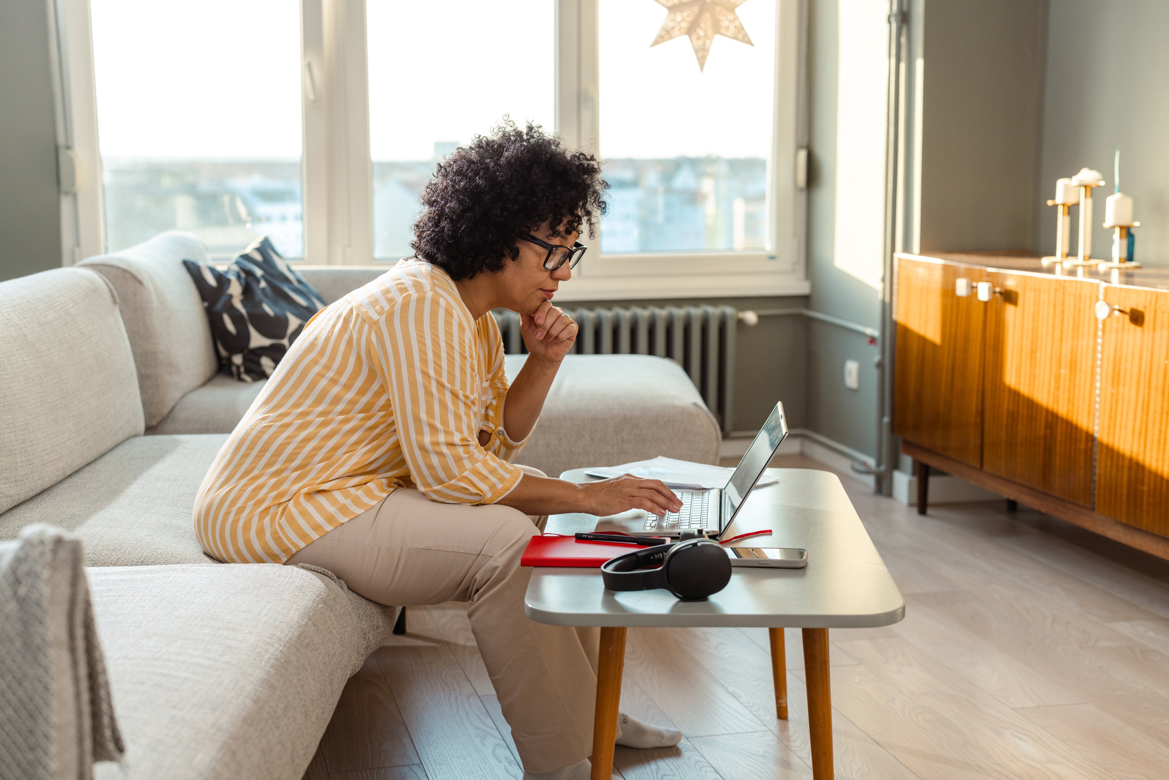 A woman sitting on the couch in a cozy living room, multitasking with a laptop and paperwork