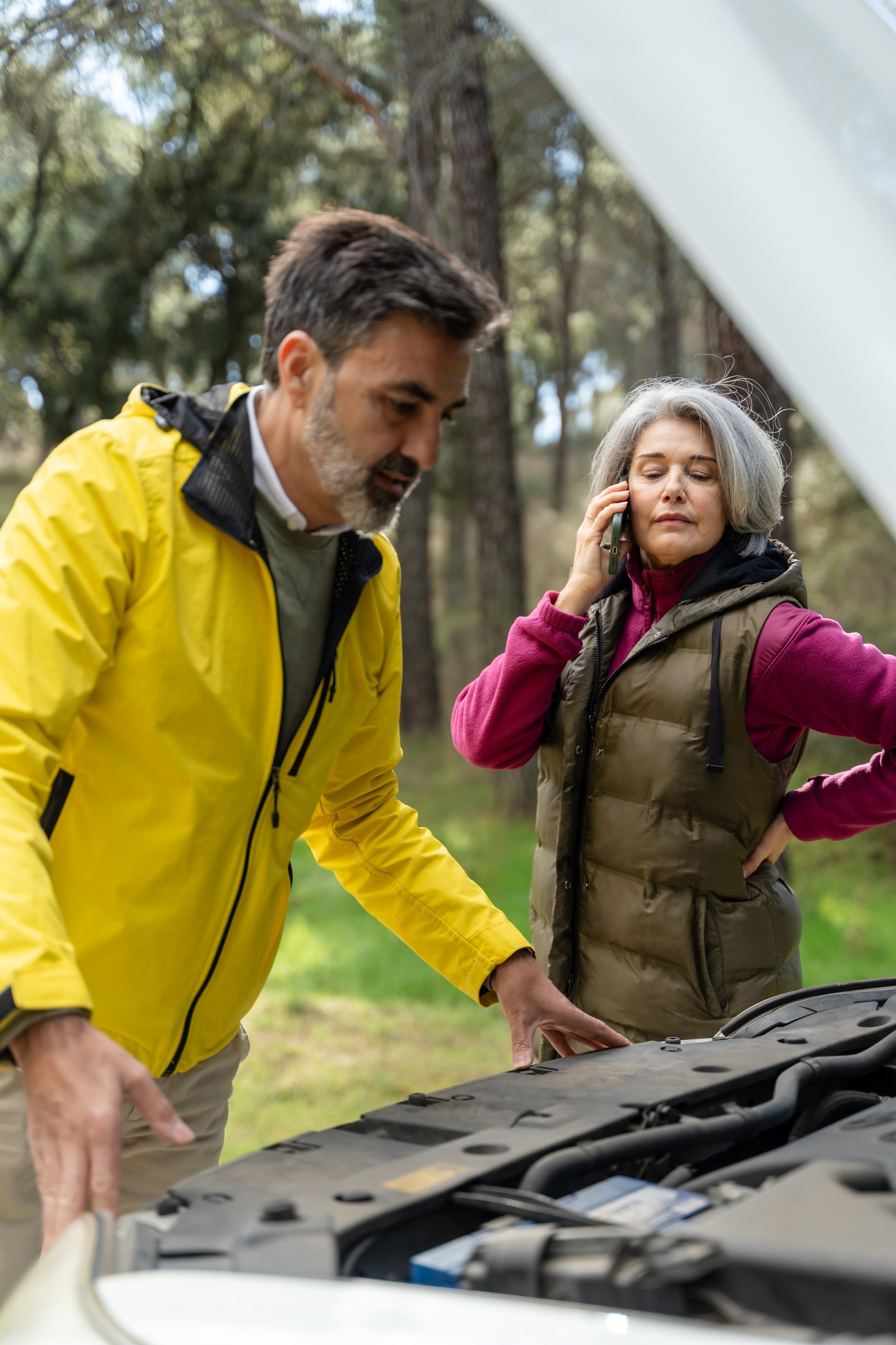 Mature couple is having car trouble in the forest, examining the engine and calling for assistance