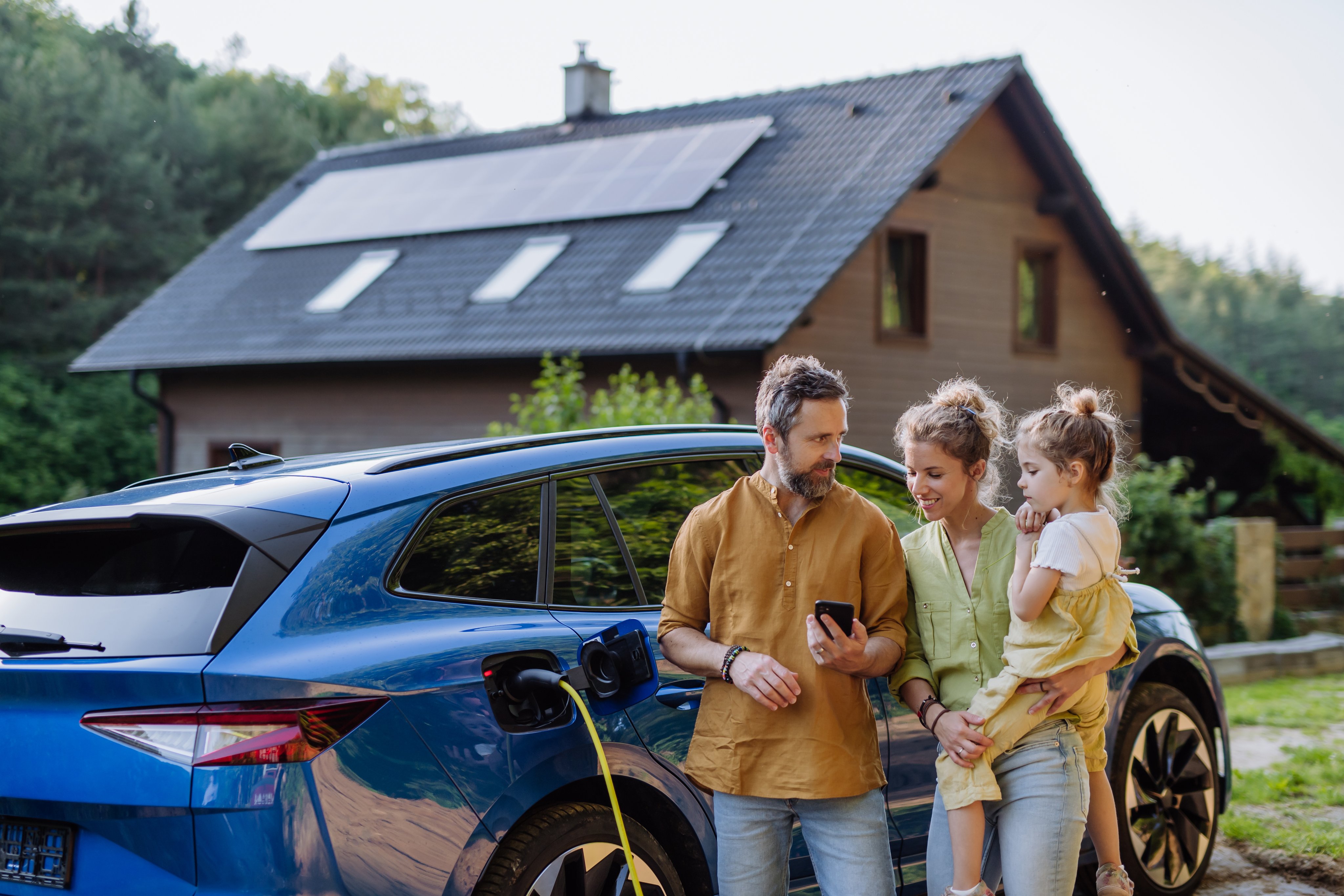Family with little daughter standing in front of their house with solar panels on the roof, having electric car