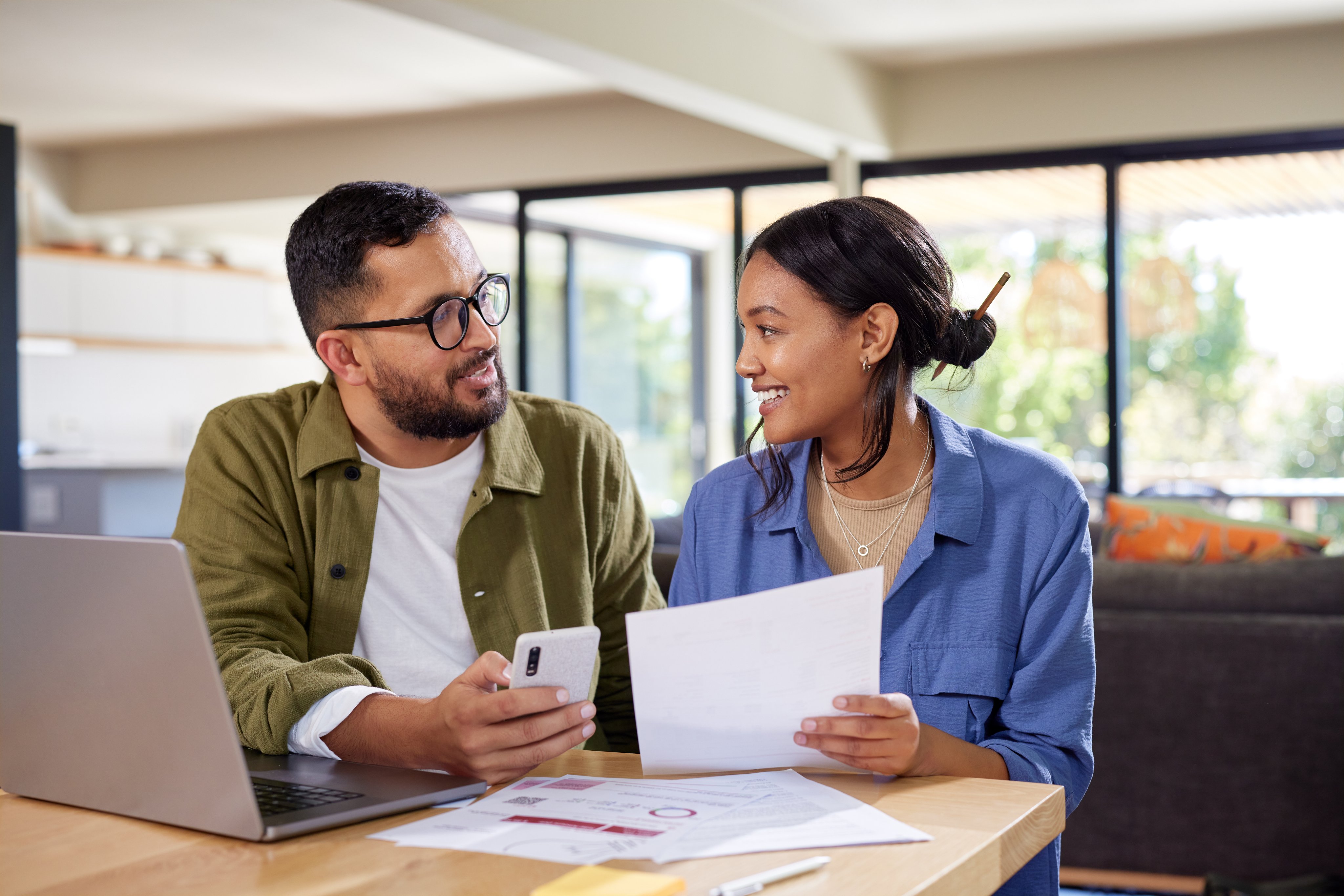 Happy indian couple discussing home finance while holding documents and working on laptop and mobile phone