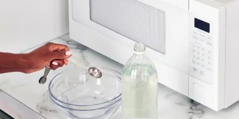 Person measuring vinegar into a bowl of water on a countertop in front of a microwave, preparing a cleaning solution