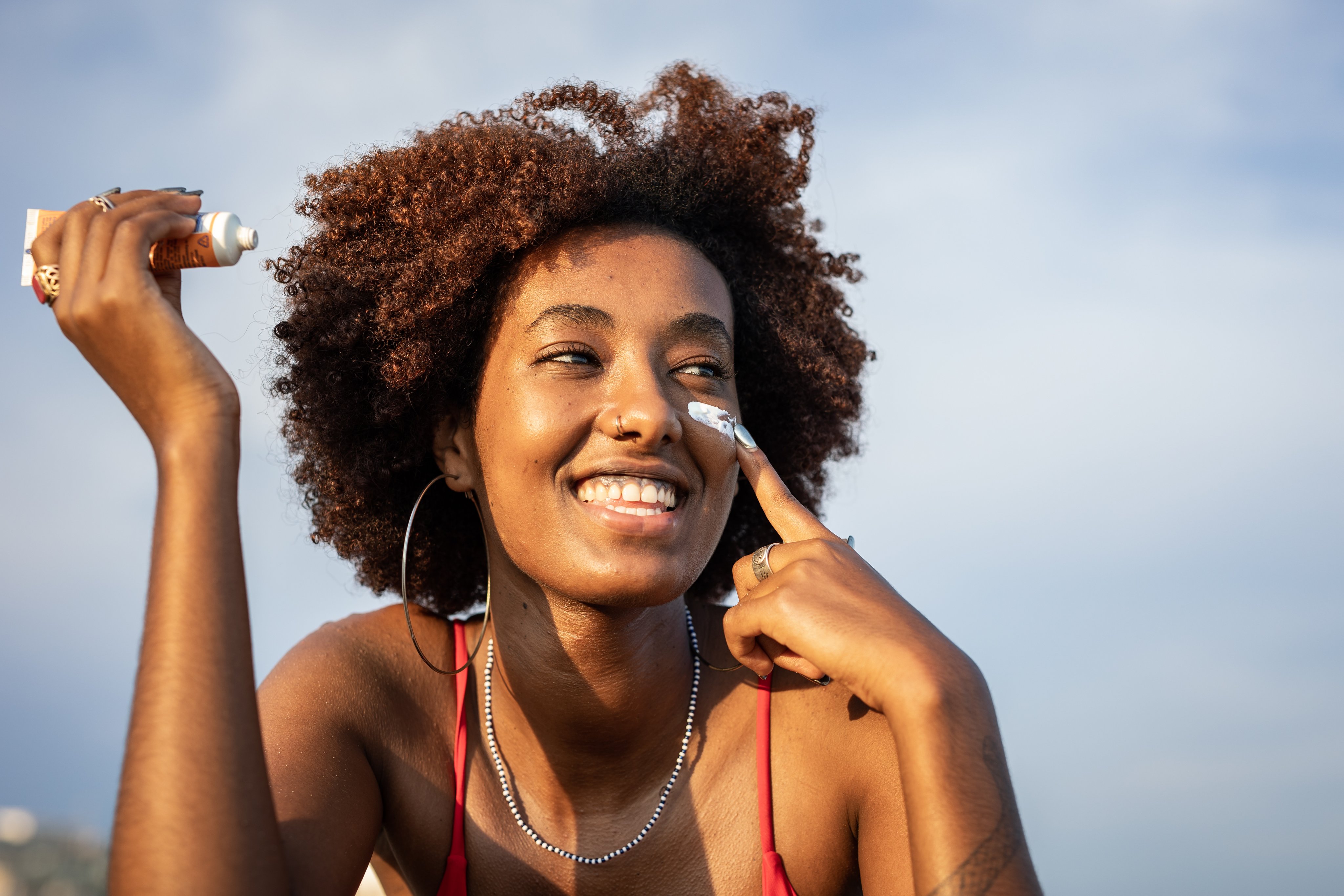 A beautiful cheerful black woman is applying sunscreen on her face.