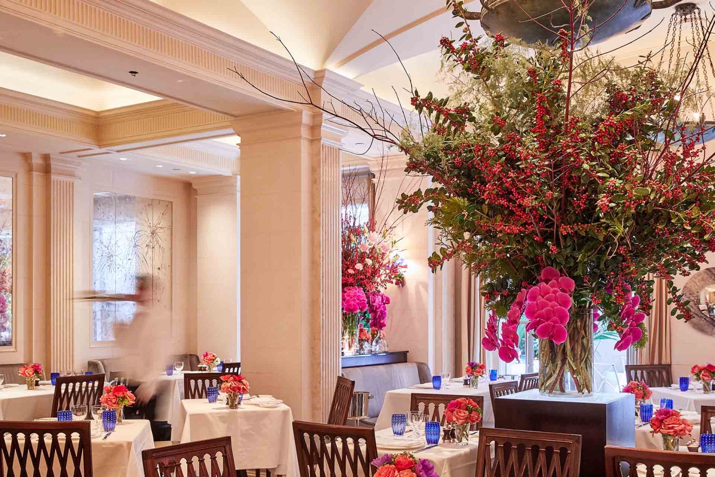 Elegant restaurant dining room with white tablecloths, wooden chairs, and vibrant floral centerpieces. Large, colorful flower arrangement is featured prominently in the foreground. Warm lighting and blurred motion of a person add ambiance.