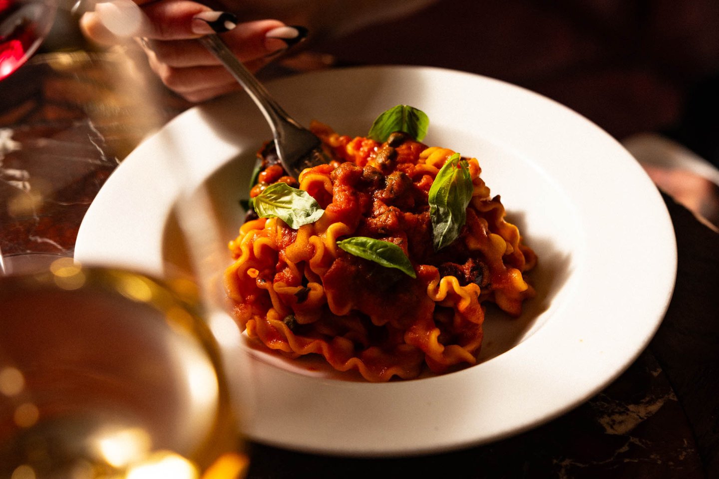 A person holds a fork over a plate of curly pasta with tomato sauce and basil leaves, set on a dark table with a glass in the foreground.