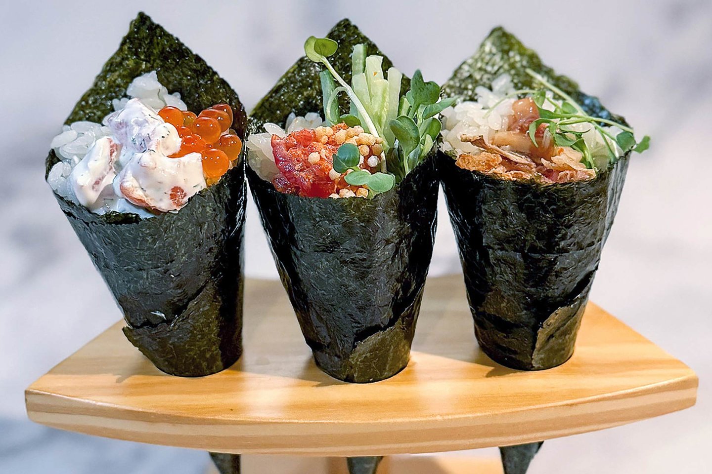 Three hand-rolled sushi cones (temaki) filled with various fresh ingredients including rice, fish, roe, and vegetables, are displayed upright on a wooden stand.