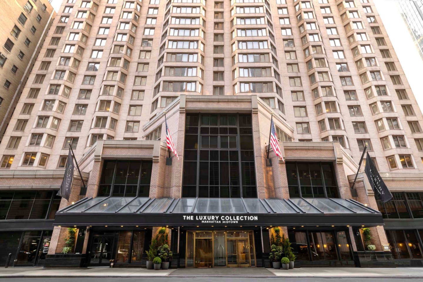 The entrance of a tall, modern hotel building with large windows, two American flags, and a sign reading "The Luxury Collection Manhattan Midtown" above glass doors. Black awnings and flags flank the entrance.