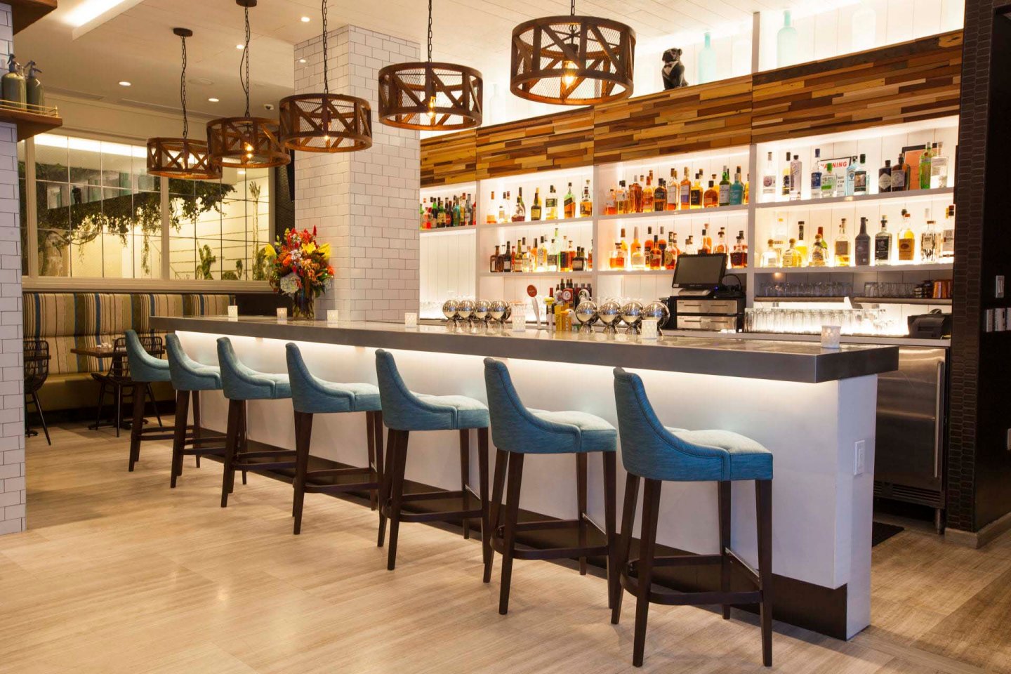 A modern bar interior featuring a row of blue cushioned stools along a sleek counter. The backlit shelves display various bottles. Pendant lights hang above, and a floral arrangement adds a touch of color to the minimalist decor.