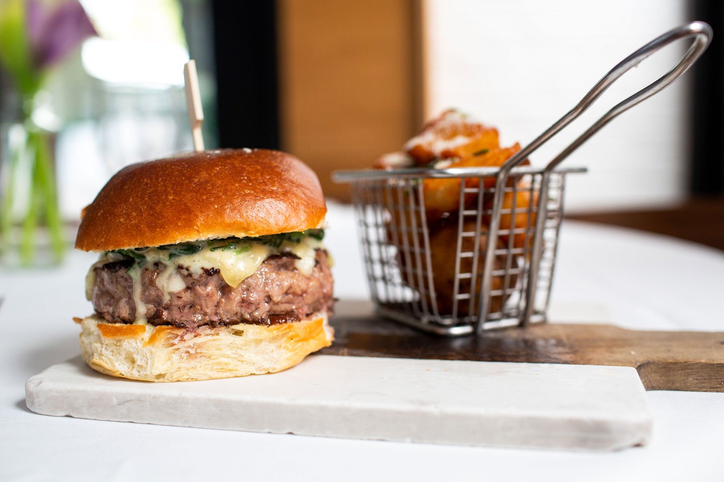 A cheeseburger with lettuce on a brioche bun sits on a marble slab, next to a metal basket filled with crispy potato wedges on a wooden board.