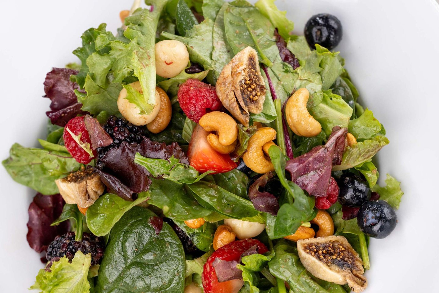 A close-up of a salad with leafy greens, cashews, dried figs, strawberries, blackberries, raspberries, and blueberries in a white bowl.