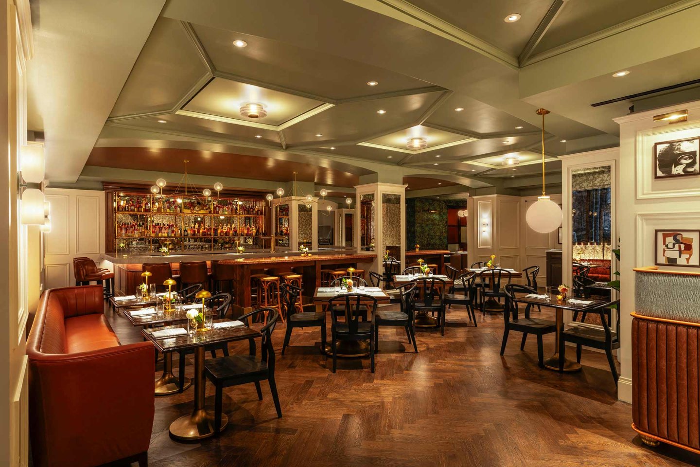 Elegant restaurant interior with wooden floors, black tables and chairs, a leather banquette, and a well-lit bar. Warm lighting and contemporary decor create a cozy, inviting atmosphere.