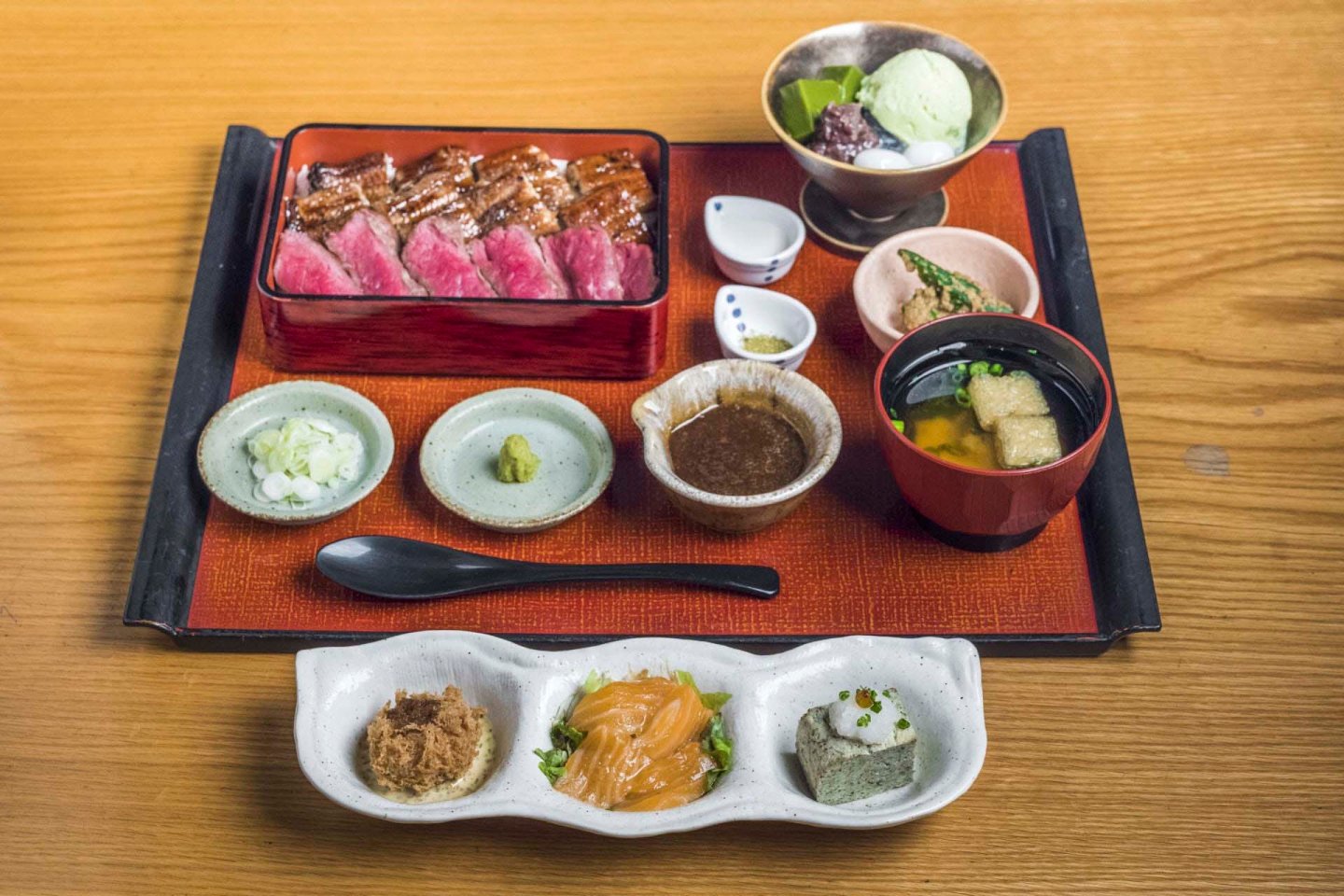 A wooden table displays a Japanese meal set including sliced steak, rice, miso soup, dipping sauces, and small dishes of pickled vegetables and tofu. A dessert with green tea ice cream is in a bowl. A small spoon rests on the tray.