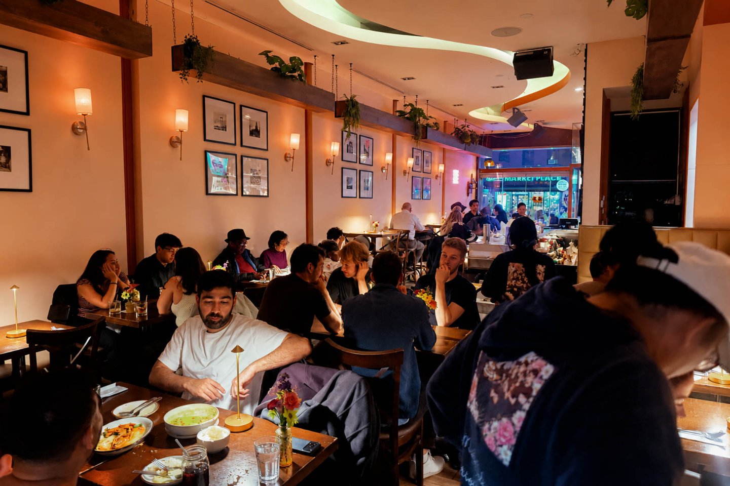 A bustling restaurant filled with people dining at wooden tables under warm lighting. Plates of food and drinks are visible, and framed photos decorate the peach-colored walls. A server attends to guests in the foreground.