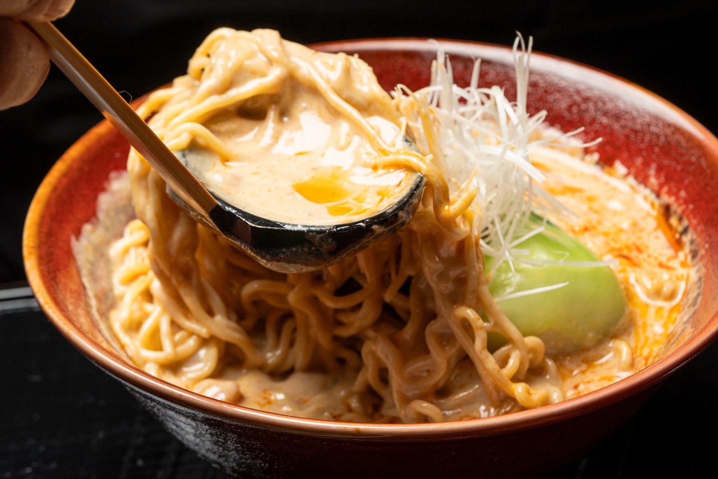 A bowl of ramen with creamy, spicy broth being poured over noodles, topped with shredded white vegetables and a green leafy garnish, served in a red bowl.