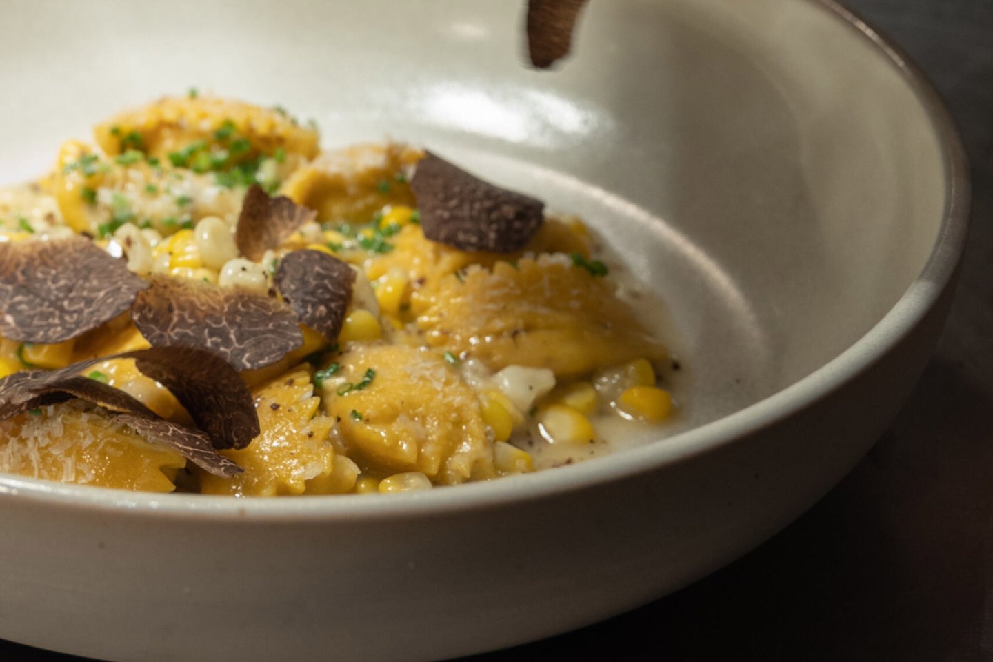 A close-up of ravioli topped with corn, shaved black truffle, and chopped chives, served in a shallow white bowl.