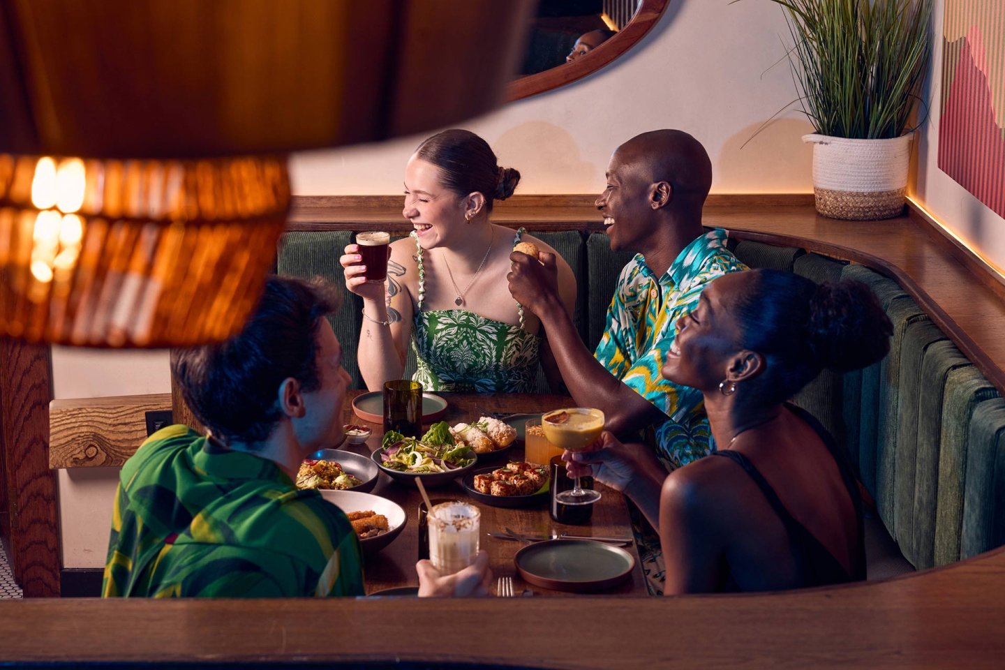 Four friends sit together in a cozy restaurant booth, laughing and enjoying cocktails and shared plates of food. The setting is warmly lit and inviting, with green decor and a relaxed atmosphere.