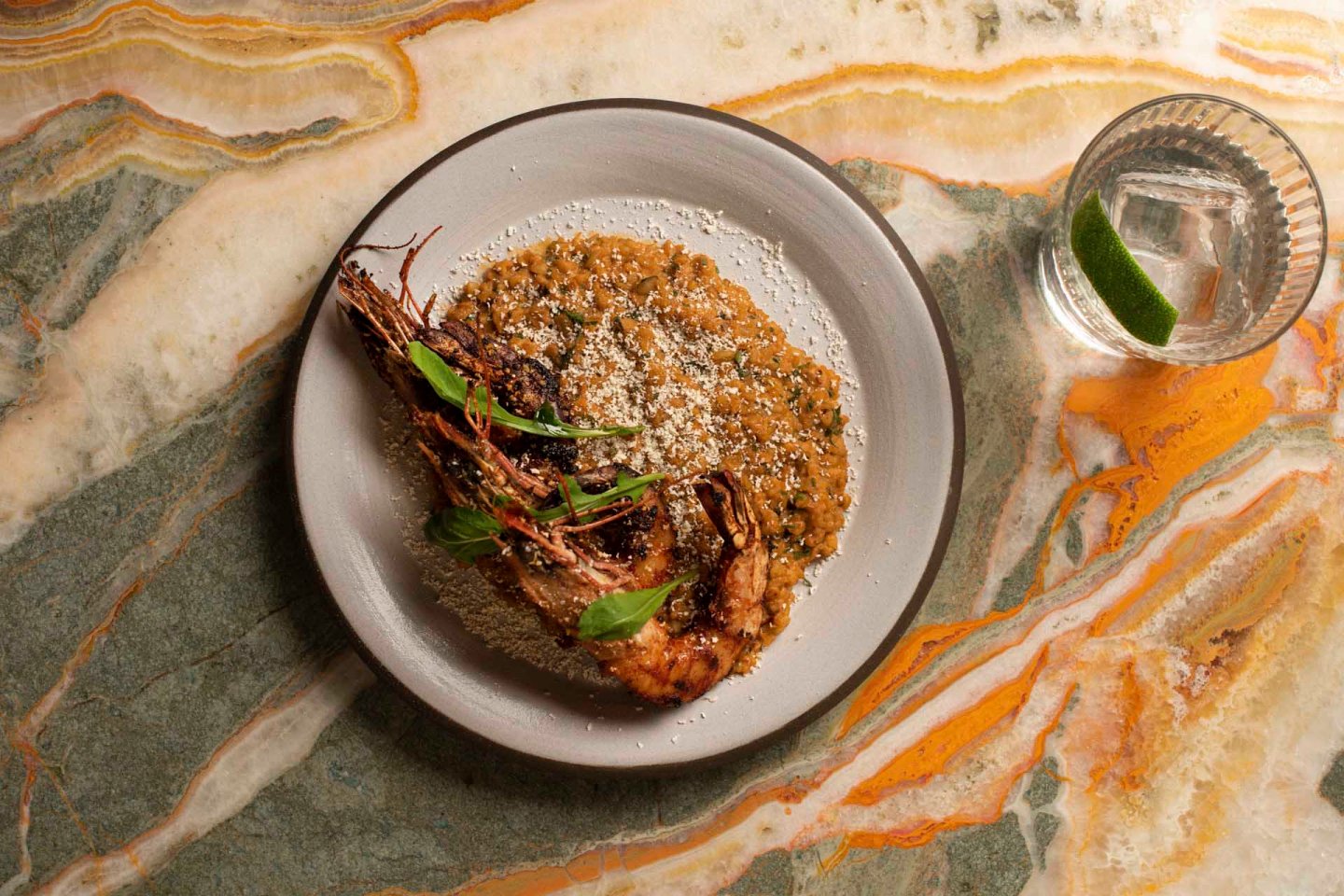 A plate of risotto topped with large shrimp and fresh greens, sprinkled with grated cheese. The dish is served on a gray plate next to a glass of clear sparkling water with a lime wedge, set on a marble surface with green, brown, and white swirls.