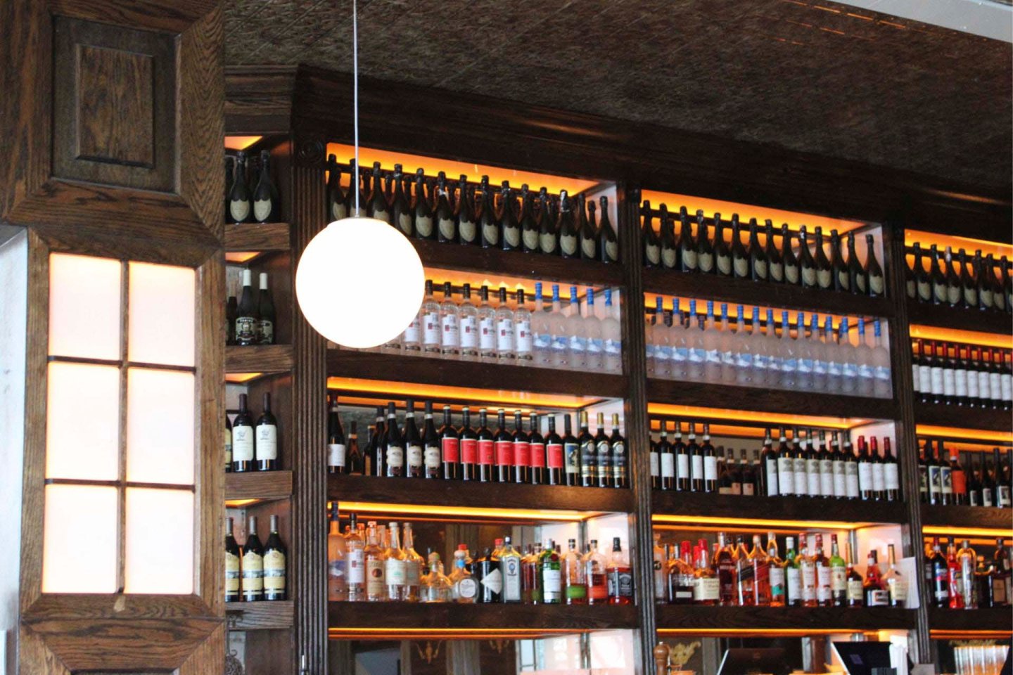 A bar with wooden shelves filled with an assortment of wine and liquor bottles. A round white light fixture hangs in the foreground, and the shelves are softly illuminated, creating a warm, inviting ambiance.