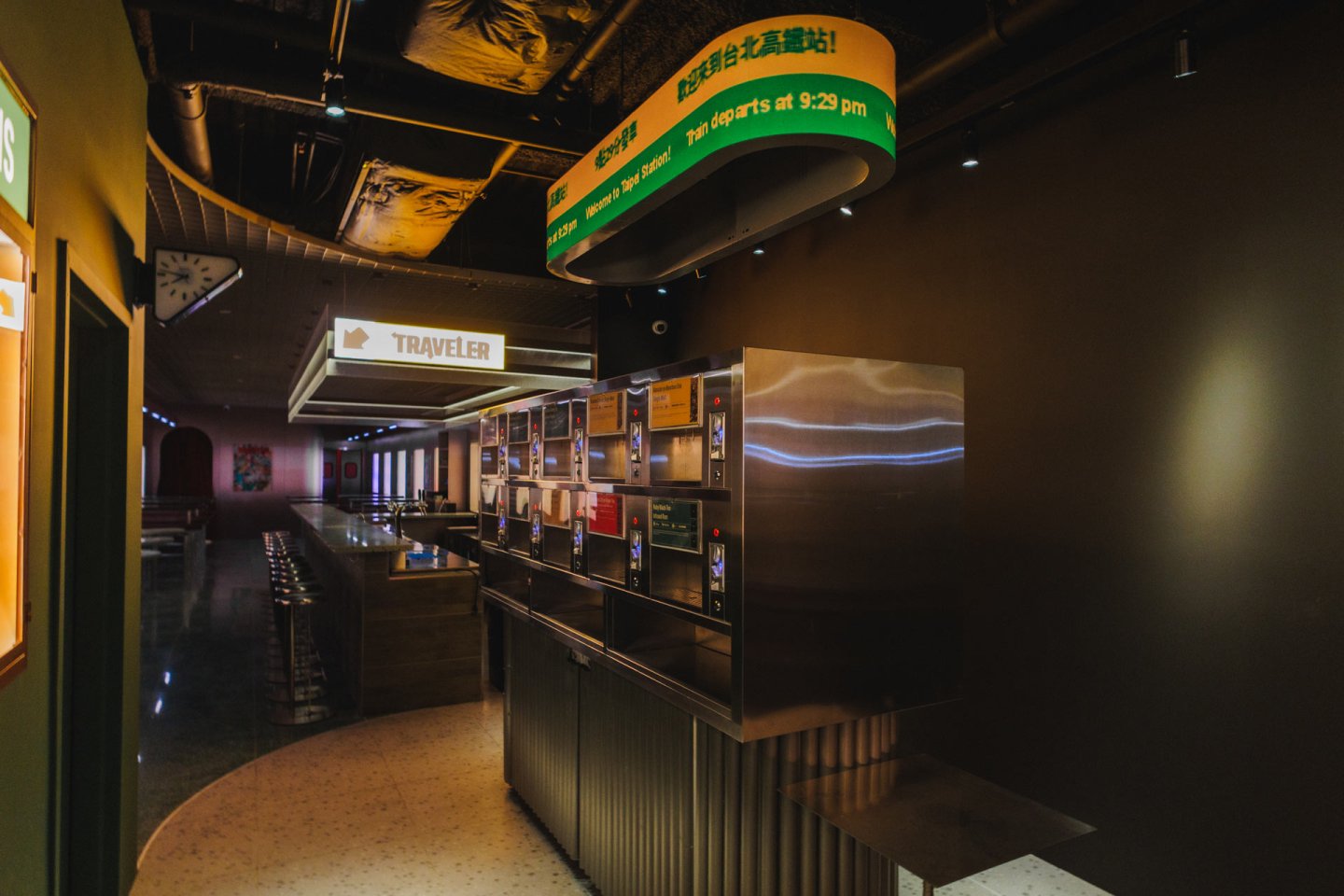A dimly lit restaurant interior with a retro-style food vending wall labeled “TRAVELER” and digital menu screens. There are rows of stools along a counter and colorful signs hanging from the ceiling.