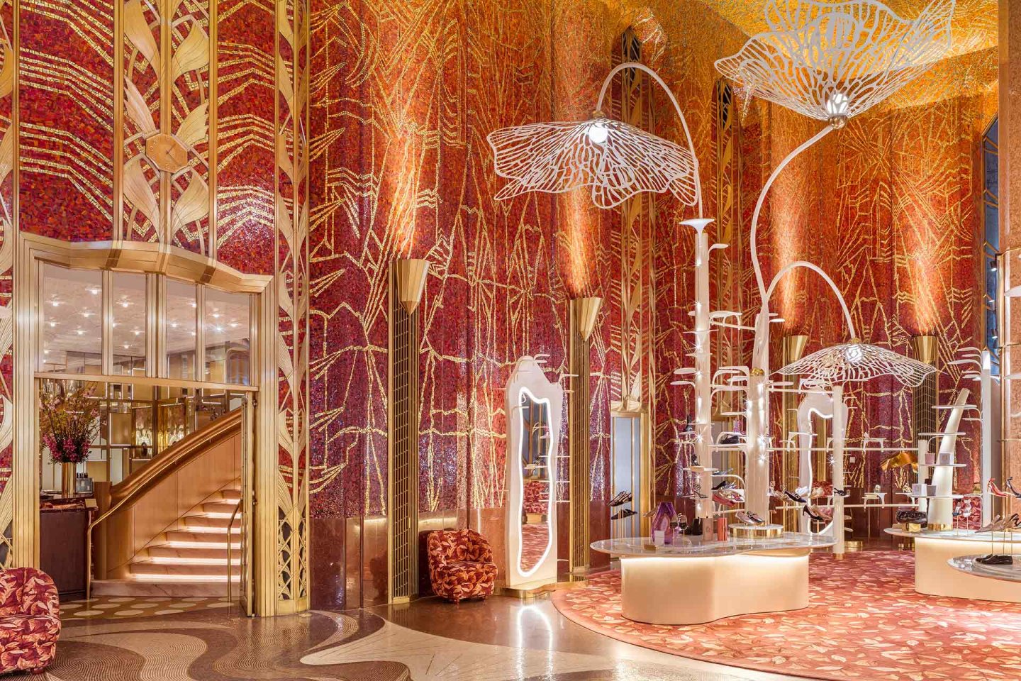 A luxurious hotel lobby features vibrant red and gold mosaic walls, tall white sculptural light fixtures, patterned carpets, plush seating, mirrored accents, and a grand staircase with glass doors.