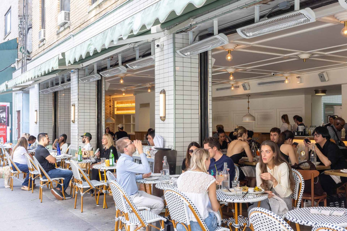 People dine and socialize at outdoor tables of a bustling café with white-tiled walls and striped chairs on a city sidewalk; some tables have drinks and food, and the café’s interior is also visible.