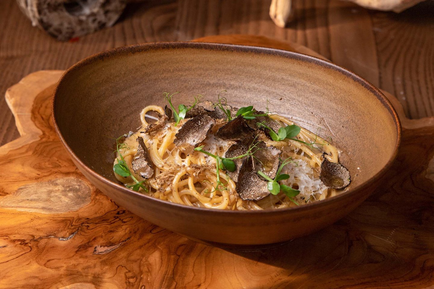 A brown bowl of pasta garnished with fresh herbs and shaved black truffles sits on a wooden surface.