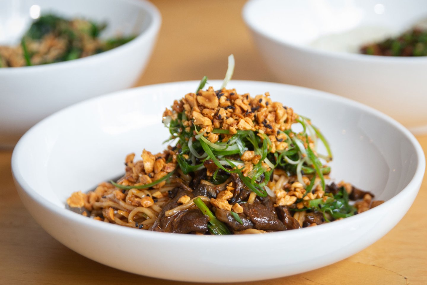 Bowl of noodles topped with chopped peanuts, green onions, and sautéed vegetables, with other bowls of food blurred in the background on a wooden surface.