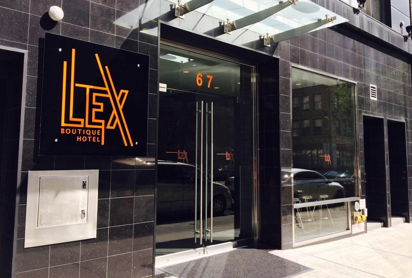 The entrance of Lex Boutique Hotel with glass double doors, a black sign with orange lettering, and the street number 67 above the door. Cars and city buildings are reflected in the glass.