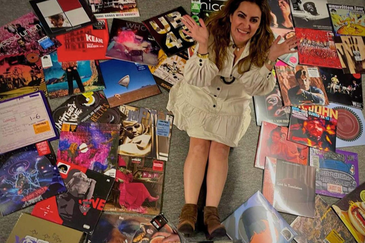 A woman in a white dress sits on the floor, smiling and holding up her hands, surrounded by numerous colorful vinyl record albums spread out in a circle around her.