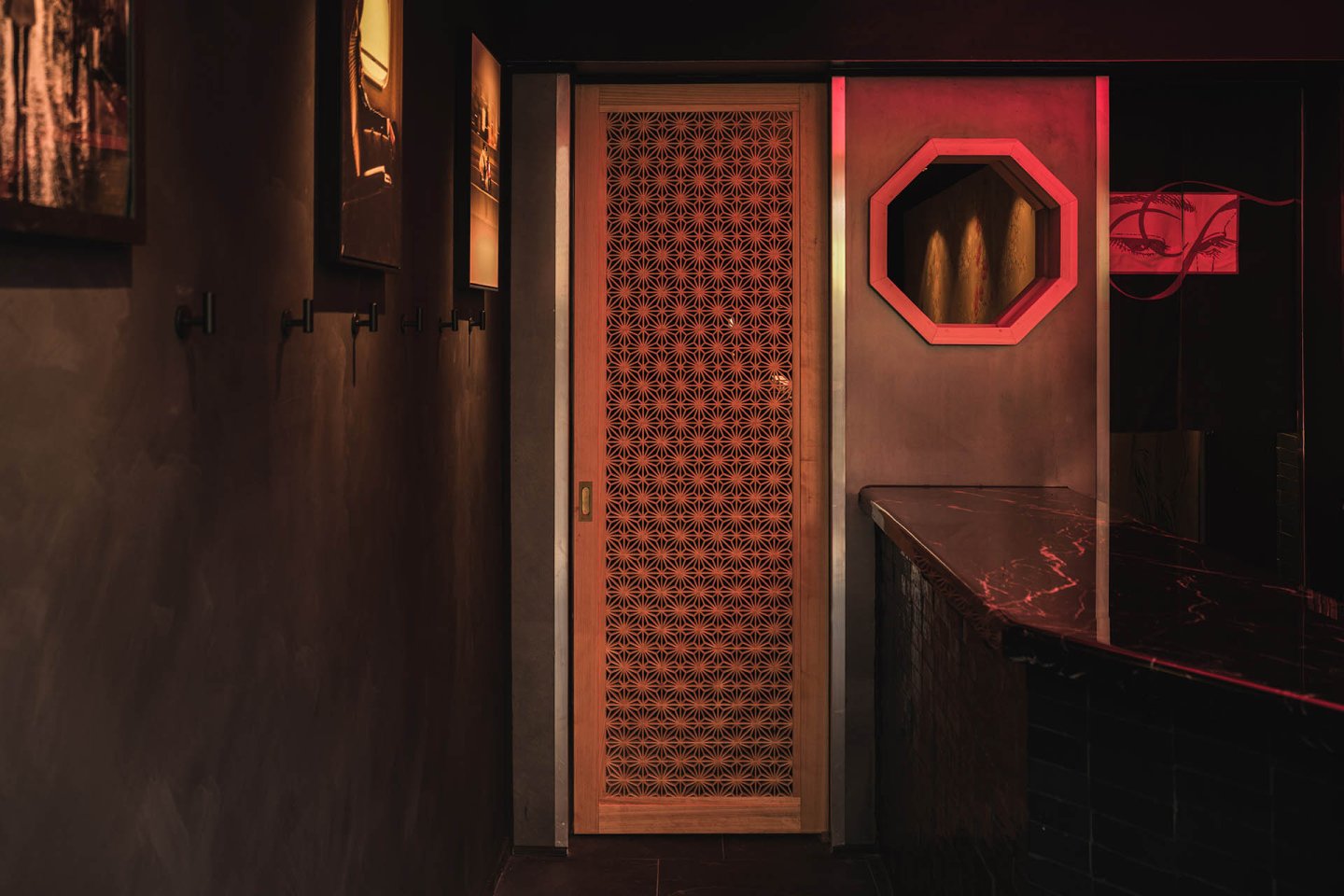 A dimly lit room with a patterned wooden door, dark walls, a black marble counter, and red lighting reflecting off a mirror and a wall decoration.
