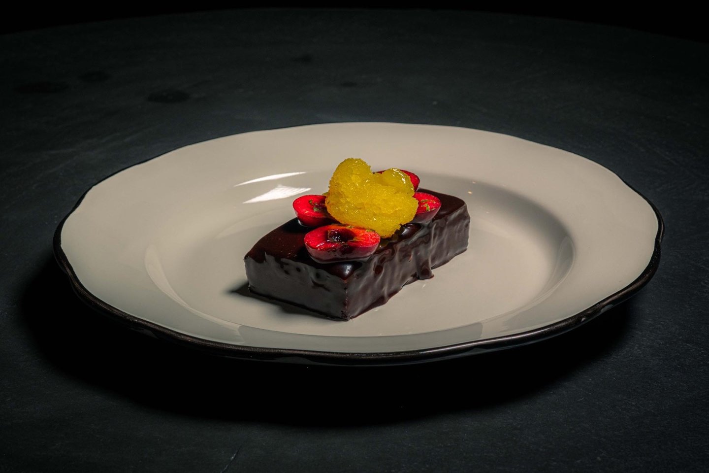 A rectangular chocolate dessert topped with cherry slices and a yellow fruit garnish sits on a white plate with a dark background.