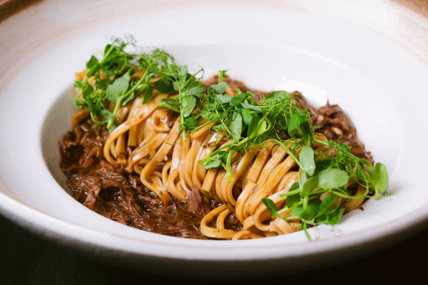 A plate of linguine pasta topped with shredded beef and garnished with fresh green herbs. The dish is arranged neatly in a white bowl.