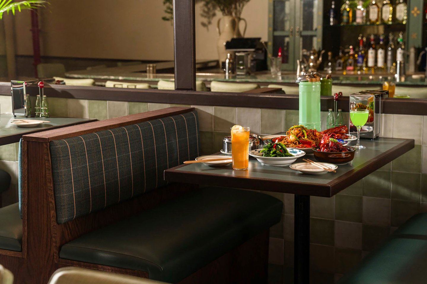 A cozy restaurant booth with green cushioned seats, featuring a table set with various dishes, including vibrant salads and meats. Two drinks are also present. The background shows a mirror and shelves with bottles.