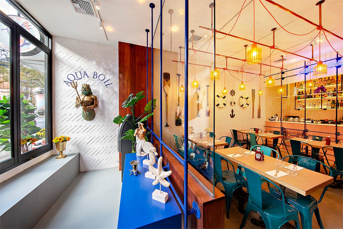 A nautical-themed restaurant interior with maritime decor, including anchors and a mermaid sculpture. Blue chairs surround a wooden table, and there's a large window with plants outside. Warm lighting creates a cozy atmosphere.