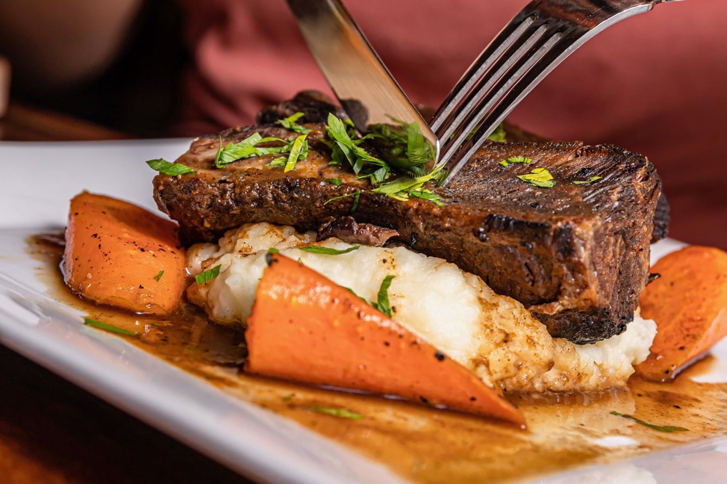 A person cuts into a piece of braised beef short rib served over mashed potatoes with roasted carrots, garnished with chopped parsley and drizzled with brown gravy on a white plate.