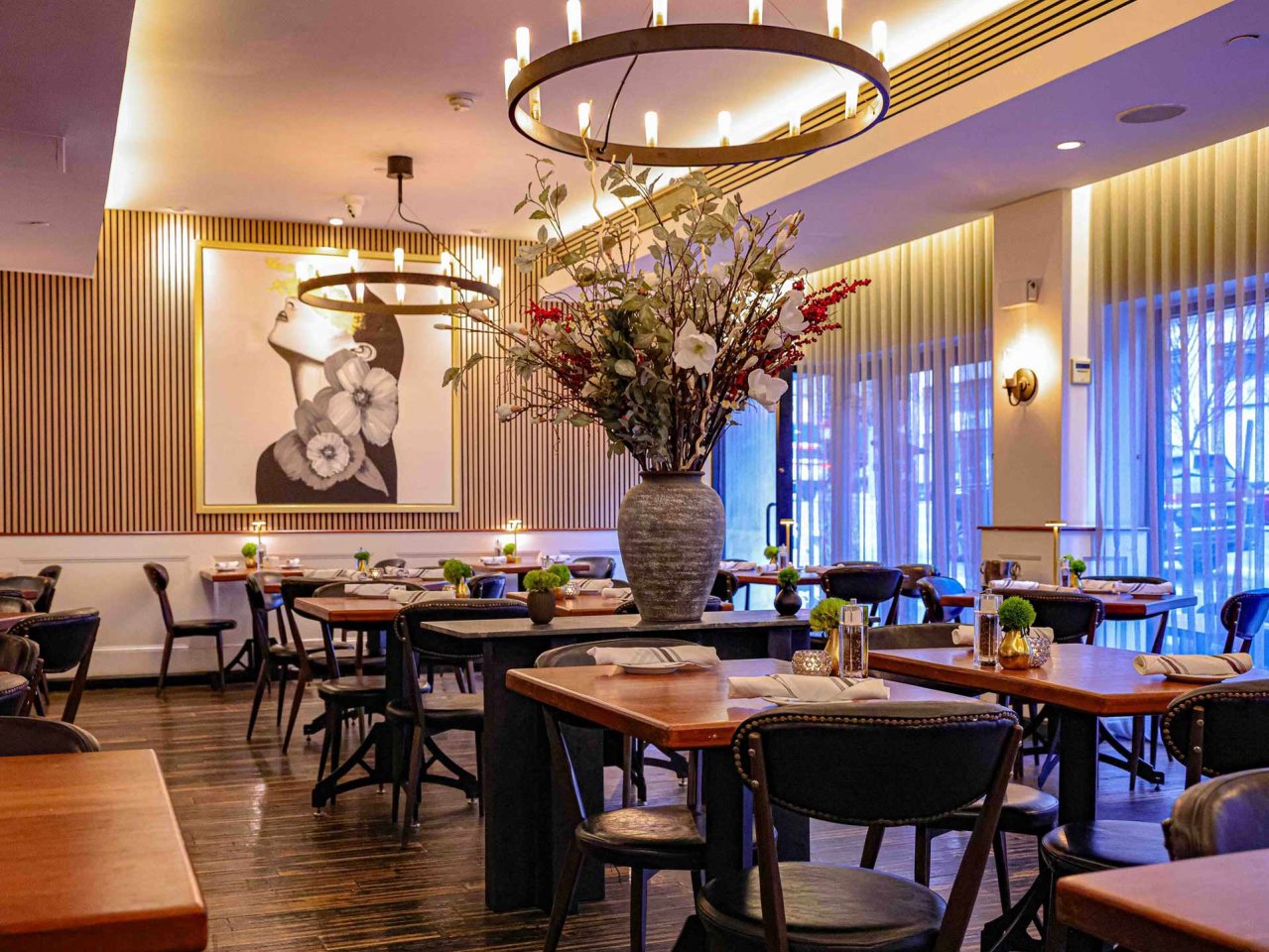 A stylish, modern restaurant interior with wooden tables, black chairs, a large floral arrangement in the center, soft lighting, and a portrait of a woman on the wall. Floor-to-ceiling windows let in natural light.