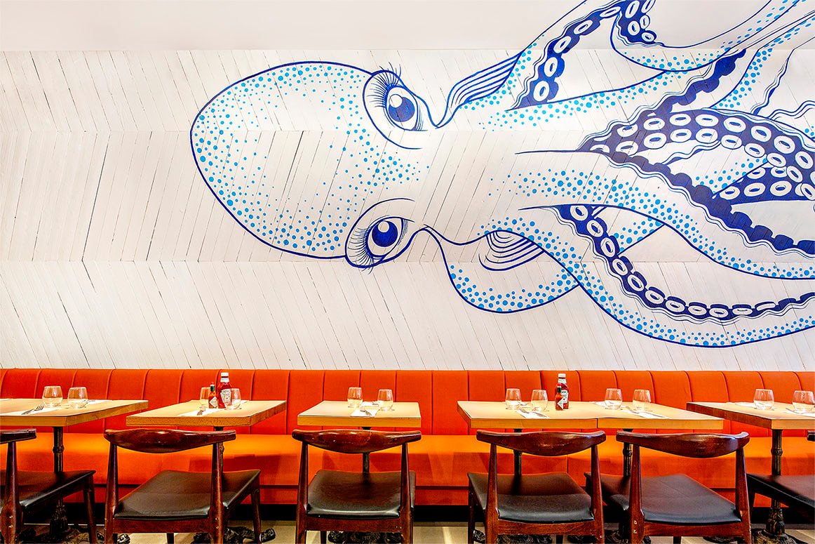 A dining area with rows of wooden tables and dark chairs against a wall featuring a large, artistic blue and white octopus mural. The tables are set with glassware and napkins, and the seating includes orange cushions.