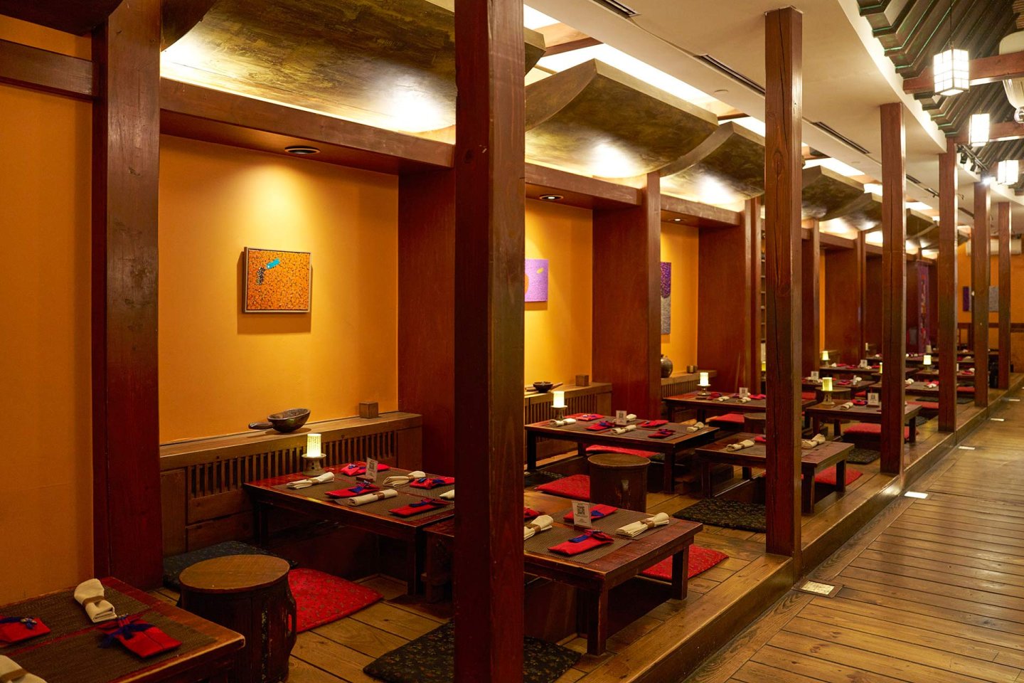 A warmly lit restaurant with wooden booths, low tables, and floor seating cushions. Each table is set with red placemats, chopsticks, and napkins. Orange walls and artwork create a cozy, inviting atmosphere.