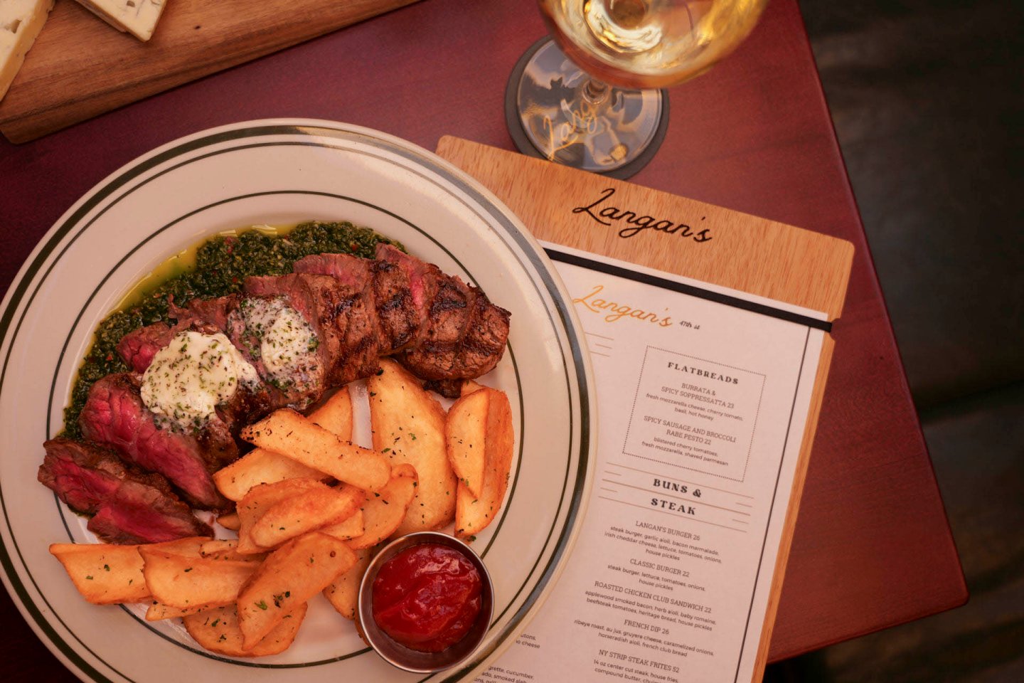 A plate with sliced steak topped with herb butter, thick-cut fries, and a side of green sauce. A small dish of ketchup is next to the fries. A glass of white wine and a menu are partially visible on the table.