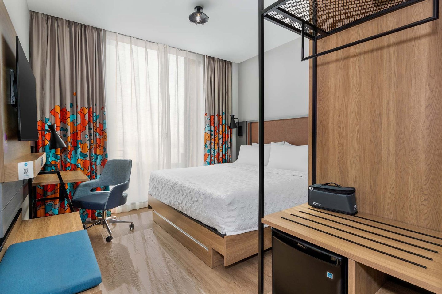 Modern hotel room with a double bed, white bedding, desk with a blue chair, wall-mounted TV, wooden bench, small fridge, and large window with patterned curtains allowing natural light in.