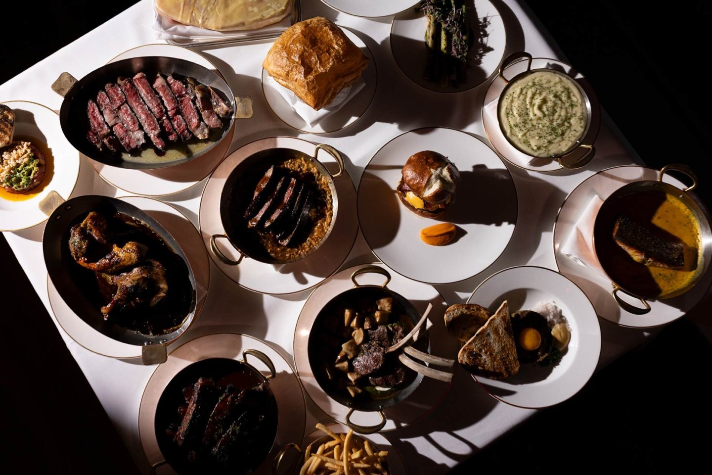 A table set with various plated meats, sauces, mashed potatoes, fries, bread, vegetables, and other sides, all presented in small serving dishes and arranged on a white tablecloth.