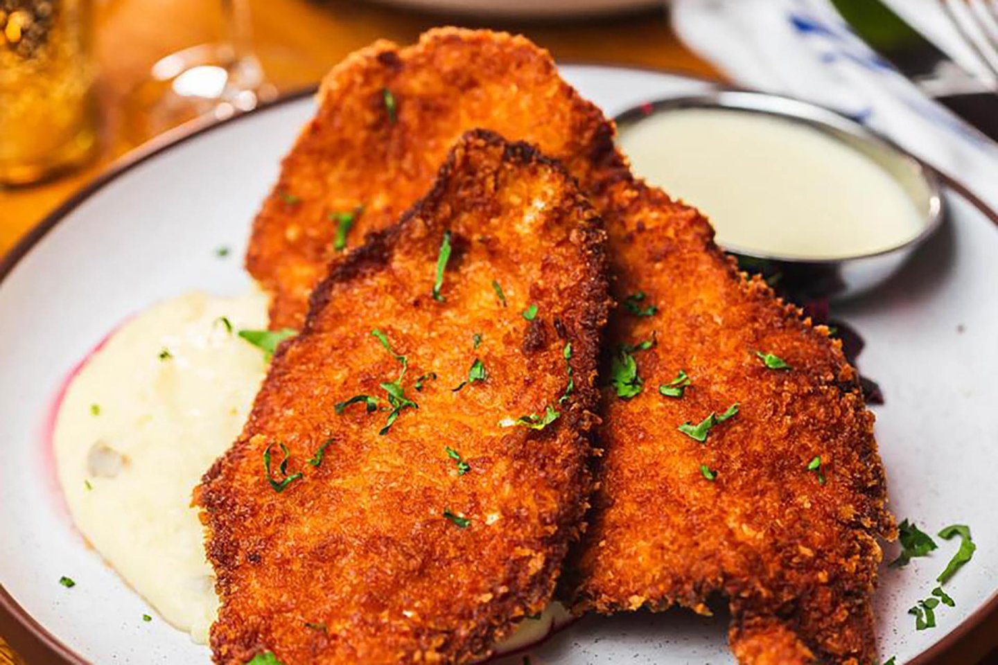 Two golden brown, breaded and fried cutlets are served on a white plate with a side of creamy mashed potatoes and a small bowl of light-colored sauce, garnished with chopped herbs.