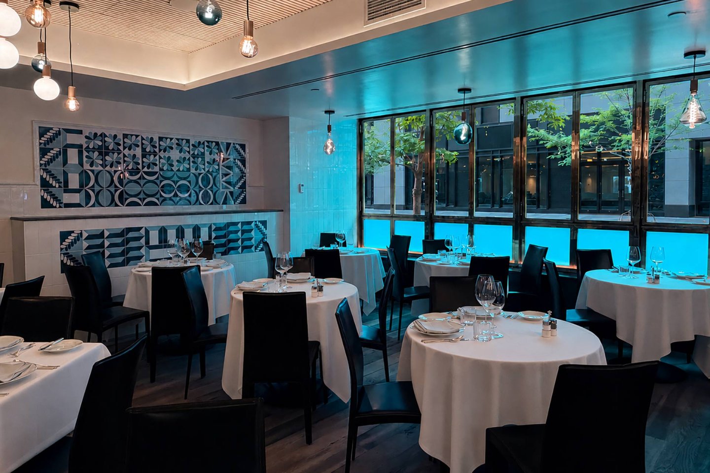 Modern restaurant interior with round tables covered in white cloths, black chairs, hanging lights, and large windows revealing trees and a blue light outside. Walls display geometric tile designs in blue and white tones.